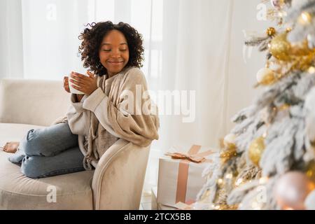 Frohe Weihnachten. Afroamerikanische Frau mit einer Tasse heißem Getränk Kaffee Tee, die neben dem Weihnachtsbaum zu Hause sitzt. Mädchen im Wohnzimmer mit Weihnachtsbaum. Weihnachtshygge Urlaub zu Hause Stockfoto