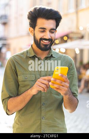 Glücklicher lächelnder bärtiger indischer Mann, der mit dem Smartphone Textnachrichten eingibt, im Internet surfen, soziale Medien, Web-App surfen, online im Freien chatten. Guy Tourist zu Fuß in der Stadt Straße. Vertikale Aufnahme Stockfoto