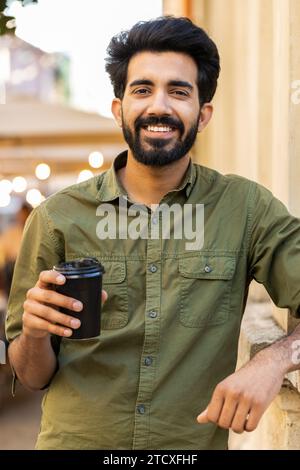 Glücklicher bärtiger indischer junger Mann, der morgens Kaffee und ein heißes Getränk genießt und den Lebensstil der Stadt im Freien genießt. Entspannen, eine Pause einlegen. Lächelnder arabischer muslimischer Erwachsener, der in der Straße des Stadtzentrums steht. Vertikal Stockfoto