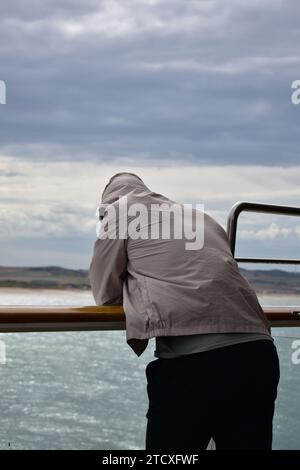 Junger Mann in schwarzer Hose und grauer Jacke mit Kappe auf einem Handlauf auf einem Schiff, das im Meer in der Nähe der Küste segelt Stockfoto