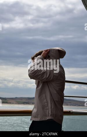 Junger Mann in schwarzer Hose und grauer Jacke, der die Kapuze seiner Regenjacke entfernt, während er neben einem Handlauf auf einem Segelschiff in der Nähe der Küste steht Stockfoto