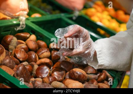 Nahaufnahme einer weiblichen Hand, die der Kamera eine Kastanie aus einem Kistenstapel im Supermarkt zeigt Stockfoto