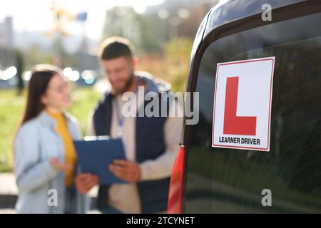 Fahrer und Kursleiter mit Klemmbrett in der Nähe des Autos im Freien, selektiver Fokus auf L-Platte. Fahrschule Stockfoto