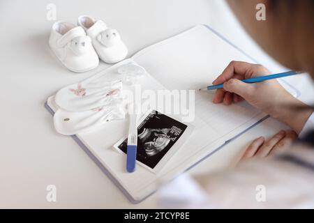 Eine schwangere Frau macht Notizen in ein leeres Notizbuch, Babyartikel und Ultraschallbilder werden platziert Stockfoto
