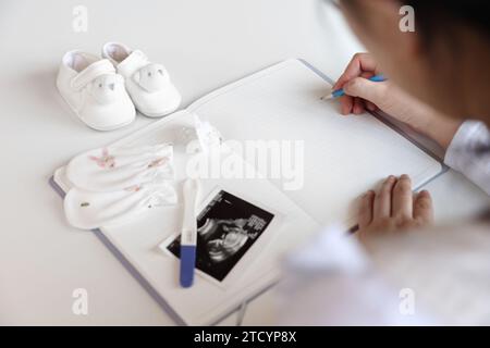 Eine schwangere Frau macht Notizen in ein leeres Notizbuch, Babyartikel und Ultraschallbilder werden platziert Stockfoto