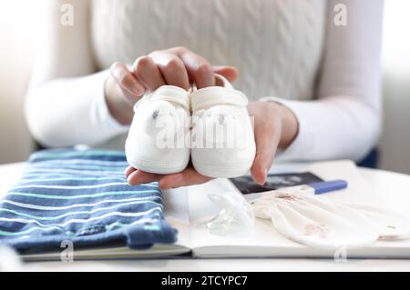 Eine schwangere Frau hält und zeigt ihre Babyschuhe, vorne sind Notizbücher und Babyartikel Stockfoto