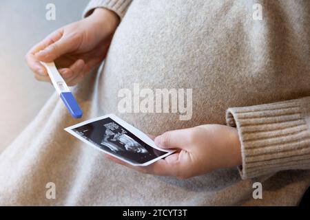 Eine schwangere Frau sitzt mit einem Ultraschallbild und einem Schwangerschaftstest Stockfoto