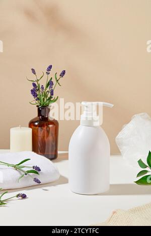 Die Vorderansicht einer unbeschrifteten Duschgel-Flasche ist auf einem Tisch mit Kerzen, Handtüchern, Lavendelblüten und einer braunen Glasvase auf einem pastellfarbenen Hintergrund zu sehen Stockfoto