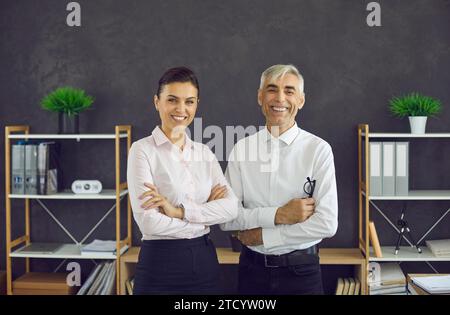 Glückliche junge Frau und ein älterer Mann stehen im Büro, schauen in die Kamera und lächeln Stockfoto