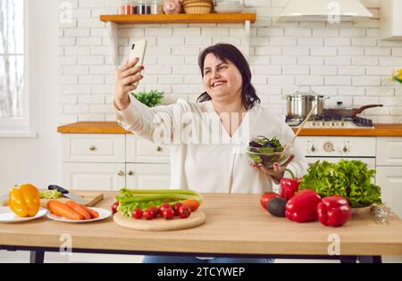Lächelnde übergewichtige Frau, die eine Schüssel mit frischem Salat hält und Selfie macht Stockfoto