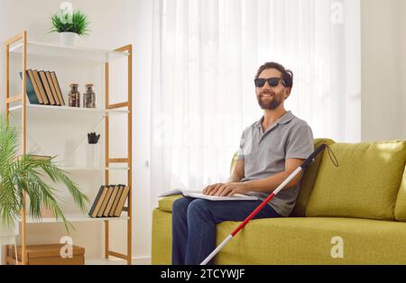 Glücklicher, sehloser Mann sitzt zu Hause auf der Couch und liest Texte in Braille-Schrift Stockfoto