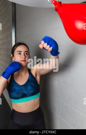 Vertikales Foto, hübsches weißes Teenager-Mädchen, brünette, in Sportkleidung und blauen Bandagen an den Händen gekleidet. Einen roten Boxball in einem Boxg schlagen Stockfoto