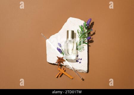 Eine unbeschriftete Parfümflasche und ein Glasstab befinden sich auf einer Steinplattform mit Lavendelblüten und Kräutern auf braunem Hintergrund. Parfümwerbung. Stockfoto