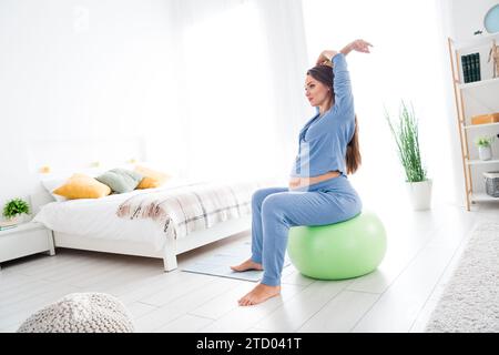 Foto eines bezaubernden, entzückenden schwangeren Mädchens, das auf Fitnessball sitzt und Yoga Meditation Aerobic ausübt und das Tageslicht drinnen ausdehnt Stockfoto