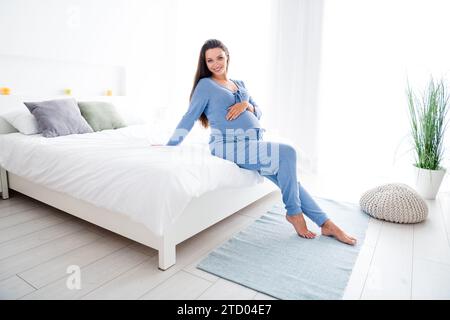 Foto von netter entzückender süßer schwangerer Mutter, die auf bequemem Bett sitzt und den Bauch berührt, der auf das Baby wartet Stockfoto