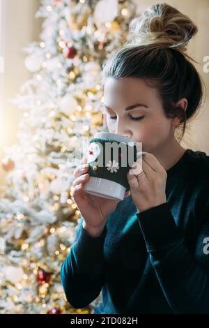Frau trinkt aus dem Weihnachtsbecher vor dem weihnachtsbaum Stockfoto