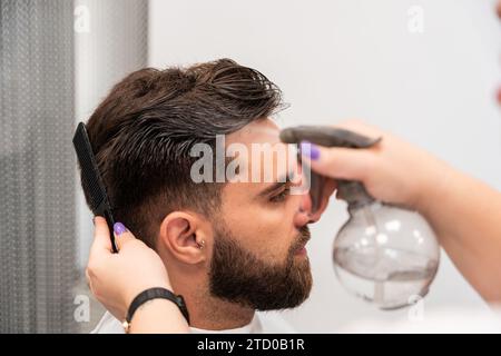 Beschneiden Hände einer nicht erkennbaren weiblichen Friseurin, die Wasser auf die Haare männlicher Kunden im Salon sprüht Stockfoto