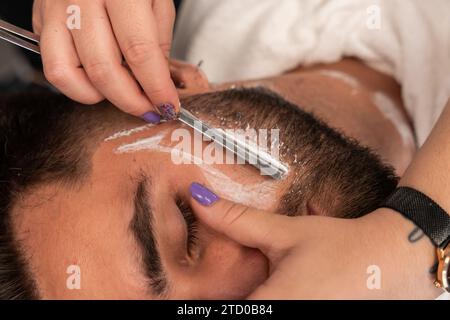 Von oben ernten Sie unerkennbare Hände von Friseurinnen, die im Salon den Bart männlicher Kunden mit Rasierer rasieren Stockfoto