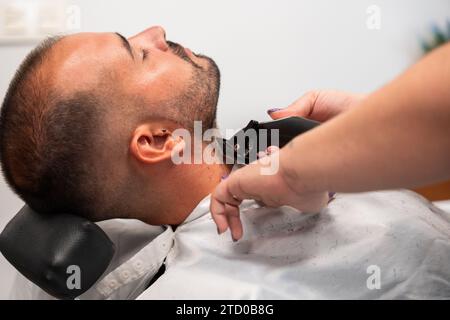 Seitenansicht der beschnittenen, nicht erkennbaren Hände eines Friseurs, der im Salon den Bart eines männlichen Kunden mit geschlossenem Auge rasiert Stockfoto