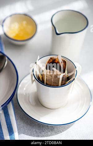 Eine heitere Szene einer tropfenden Kaffeetasse mit Filter, die auf einem Teller neben einem silbernen Krug und einer Schüssel Honig steht und in sanftes Morgenlicht getaucht ist. Stockfoto