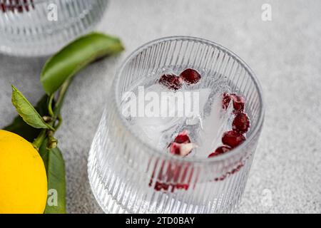 Nahaufnahme eines Gin Tonic Glases mit Eiswürfeln, garniert mit Zitronen- und Granatapfelkernen, serviert auf einer strukturierten grauen Oberfläche. Stockfoto
