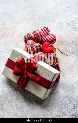 Eine Geschenkbox, die mit einem roten Band gebunden ist und mit verschiedenen handgefertigten Herzornamenten vor einem strukturierten Hintergrund verziert ist. Stockfoto