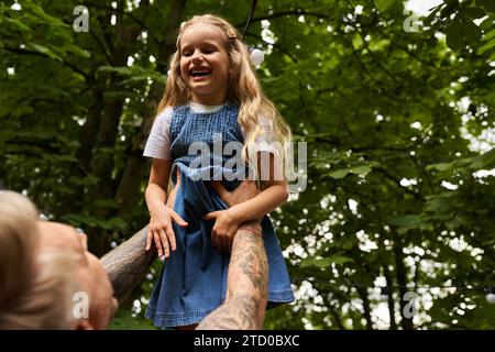 Überglücktes Mädchen lacht in den Händen des tätowierten Vaters im grünen Park, Freizeit und spielt zusammen Stockfoto