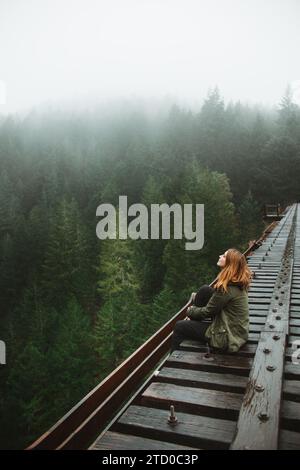 Eine nachdenkliche Frau sitzt auf einer Eisenbahnstrecke inmitten der nebeligen immergrünen Wälder von Vancouver Island, die sich in Einsamkeit widerspiegelt. Stockfoto