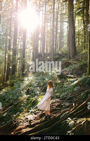 Ein junges Mädchen in hellem Kleid wandert durch einen dichten, sonnendurchfluteten Wald auf Avatar Grove, Vancouver Island und verkörpert ein Gefühl von Abenteuer und Ruhe Stockfoto