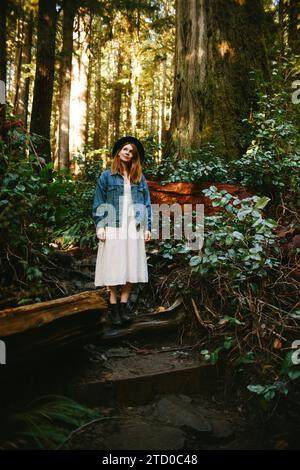 Eine junge Frau in lässigem Outfit und Hut steht auf einem Waldweg umgeben von hohen Bäumen auf Avatar Grove, Vancouver Island. Stockfoto