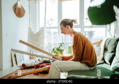 Seitenansicht einer jungen Frau in legeren Kleidern, die auf dem Sofa sitzt und zu Hause die Verpackung für Weihnachtsgeschenke in der Hand hält Stockfoto