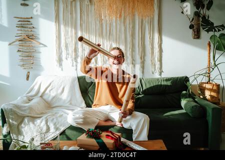 Lächelnde junge Frau, die auf einem bequemen Sofa sitzt und Weihnachtsgeschenke hält, während sie zu Hause Spaß hat Stockfoto