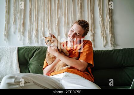 Junge Frau, die die entzückende britische Kurzhaar-goldene Katze umschließt, während sie auf einer bequemen Couch vor dem verschwommenen Innenraum des Wohnzimmers zu Hause sitzt Stockfoto