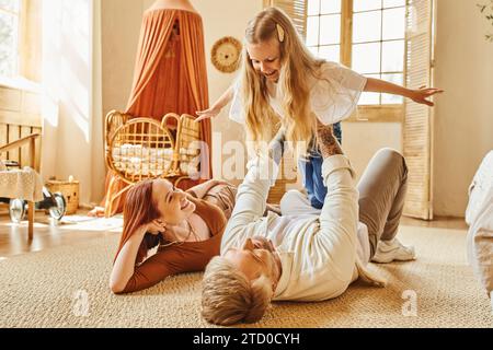 Fröhliche Frau, die den Mann ansieht, der mit der Tochter auf dem Boden im Wohnzimmer spielt, bindende Momente Stockfoto