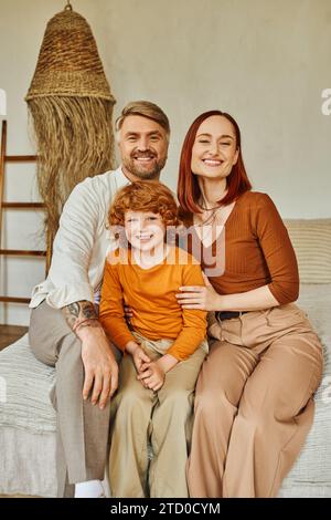 Fröhliche Eltern mit rothaarigem Sohn, der auf dem Bett sitzt und in gemütlicher Atmosphäre in die Kamera blickt Stockfoto