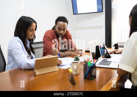Ernsthafte Geschäftsleute untersuchen Dokumente und verwenden ein Tablet, während Kollegen während der Besprechung im Büro über Laptop im Hintergrund diskutieren Stockfoto