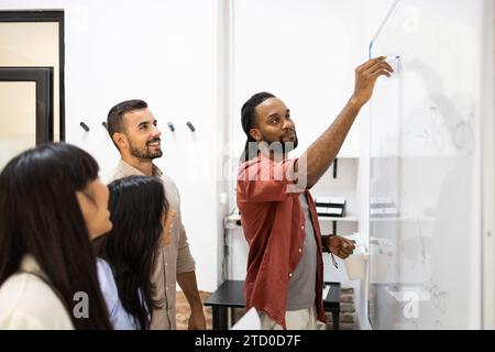 Lächelnder Geschäftsmann, der auf Whiteboard schreibt und männlichen und weiblichen Kollegen während eines Geschäftstreffens im Büro erklärt Stockfoto