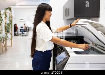 Seitenansicht einer lächelnden asiatischen jungen Geschäftsfrau in Casuals mit langem Haar mit modernem Kopierer während der Arbeit im kreativen Büro Stockfoto