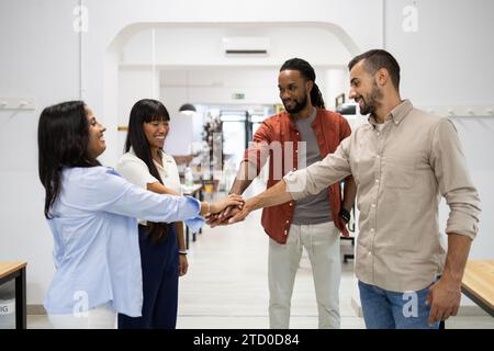 Fröhliche, multirassische männliche und weibliche Kollegen, gekleidet in Casuals, die Hände stapeln, während sie im kreativen Büro stehen Stockfoto