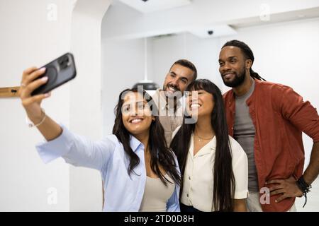 Lächelnde junge Geschäftsfrau macht Selfie mit fröhlichen Kollegen aus verschiedenen Rassen über das Smartphone, während sie im kreativen Büro steht Stockfoto