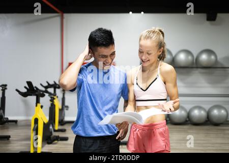 Eine junge Kundin bespricht ihr neues Trainingsprogramm mit einem männlichen Fitnesstrainer in einem Fitnessstudio, mit Trainingsgeräten im Hintergrund Stockfoto