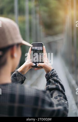 Sehen Sie anonyme Wanderer mit Mütze und Gelegenheiten, die beim Wandern im Wald am Wochenende die Brücke mit dem Smartphone fotografieren Stockfoto