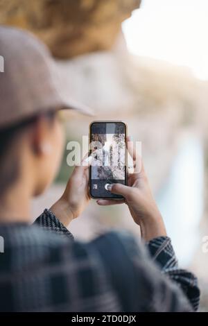 Sehen Sie anonyme Wanderer mit Mütze und Gelegenheiten, die beim Wandern im Wald am Wochenende die Brücke mit dem Smartphone fotografieren Stockfoto