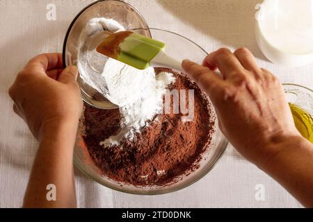 Draufsicht auf die Hände einer anonymen Person gießen Sie sorgfältig gesiebtes Mehl in eine Schüssel mit Kakaopulver für ein Backrezept vor weißem Hintergrund Stockfoto