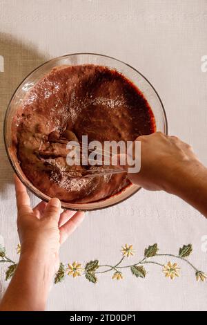 Blick von oben auf die Hände einer anonymen Person, die Schokoladenteig in einer klaren Glasschale auf einem floralen gestickten Tuch rastet Stockfoto