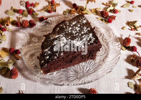 Hoher Winkel einer Scheibe reichhaltigen Schokoladenkuchen, mit Puderzucker auf einer dekorativen Glasplatte, umgeben von getrockneten Blumen Stockfoto