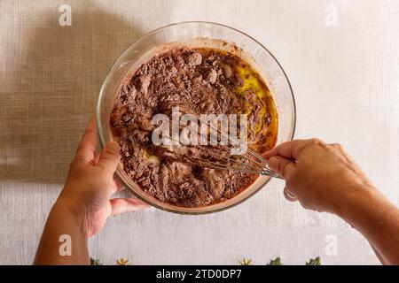 Draufsicht auf die Hände einer anonymen Person, die mit einem Schneebesen Schokoladenteig in einer klaren Glasschale auf einem Küchentisch mischt Stockfoto
