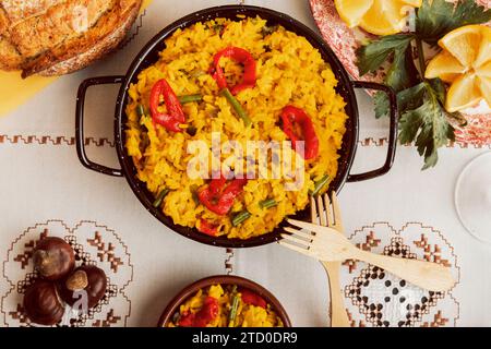Paella in traditioneller Pfanne mit Zitronen- und Tomatengarnitur, umgeben von verschiedenen Gerichten. Stockfoto