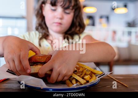 Nahaufnahme eines hungrigen Elementarmädchens mit köstlichem, appetitlichem Hamburger serviert mit pommes Frites auf Teller auf Holztisch im Restaurant Stockfoto