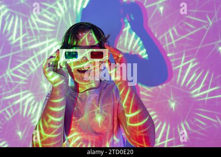 Porträt eines lächelnden Mädchens in lässiger Kleidung und einer 3D-Brille mit Perlen im Gesicht, die in beleuchteten Neonlichtern im Studio mit offenem Mund und geschlossenem Mund steht Stockfoto